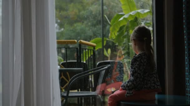Маленька дівчинка сидить і дивиться на задній двір зсередини. Хоче грати, але його сильний дощ надворі — стокове відео