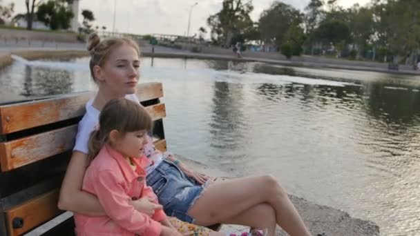 Madre joven con niña disfrutando de la tarde soleada en el parque cerca del lago. Sentado en el banco . — Vídeos de Stock