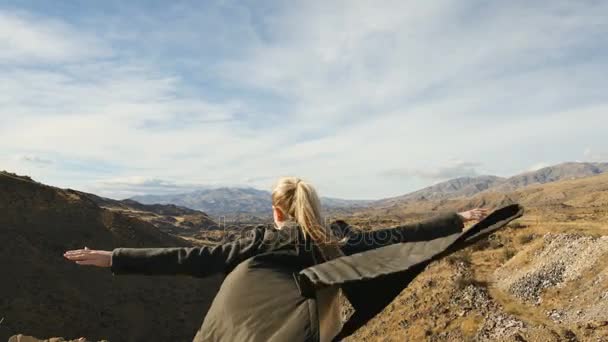Mujer joven de pie en el borde del acantilado y levantando las manos contra las altas montañas rocosas tomando el sol en el sol de la noche. Chica feliz disfrutando de éxito y vista impresionante . — Vídeo de stock