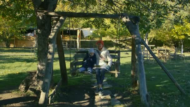 Grandmother and child ride on the wooden swing in the park outdoor in the morning. — Stock Video