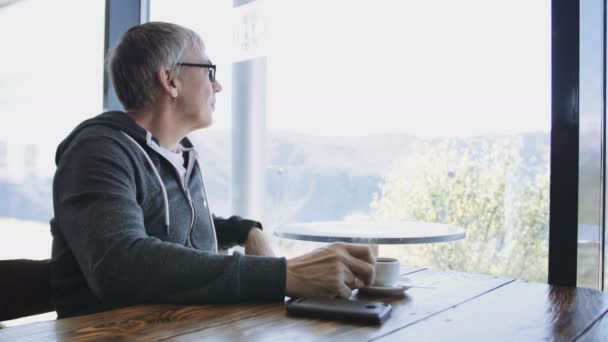 Senior kaukasiska sport stil man tittar ut genom fönstret på café. År grå mannen njuter dricka kaffe vid bordet med mobiltelefon — Stockvideo