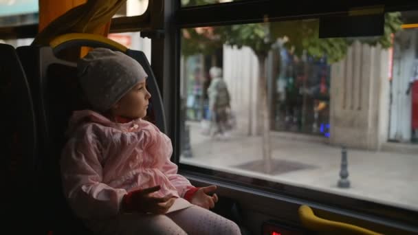 Niña rubia con una chaqueta rosa, Kid está sentado en una cabina de autobús. Sentada en su asiento y mirando a través de la ventana de vidrio . — Vídeos de Stock
