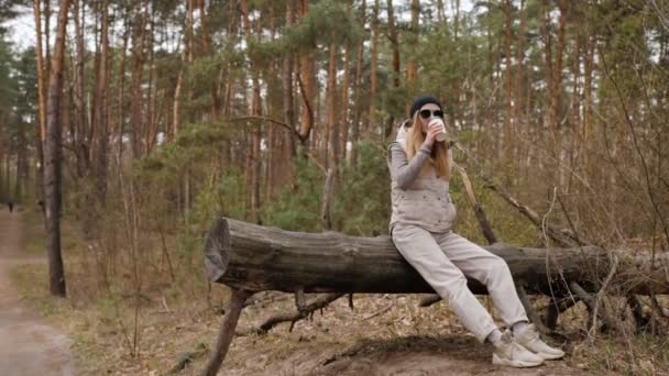 Ampla para fechar tiro jovem bela mulher loira em colete e óculos de sol sentado em log em um fundo da floresta com xícara de papel de café — Vídeo de Stock