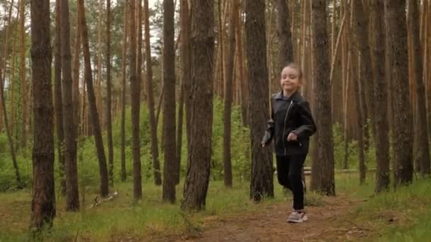 The little girl runs along the path road in the pine forest. Dressed in the leather jacket. — Stock Video