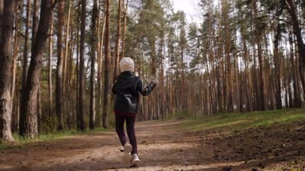 The little girl runs along the path road in the pine forest. Dressed in the leather jacket. — Stock Video