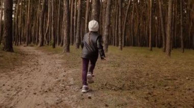 Küçük kız çam ormanında yol boyunca koşar. Deri ceket giymişti..