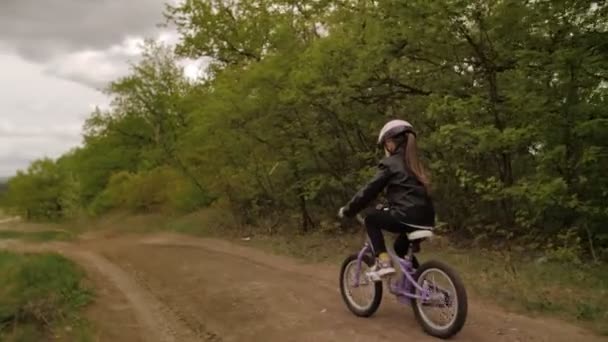 Söt liten flicka i hjälm rida en cykel på landsvägen mellan vit kulle och skog. Steadycam slow motion shot — Stockvideo