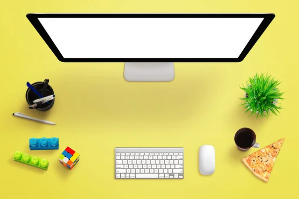 Yellow desk with computer display, keyboard, mouse, tea, coffee, plant, pizza, cubes and pens. Free space for text.