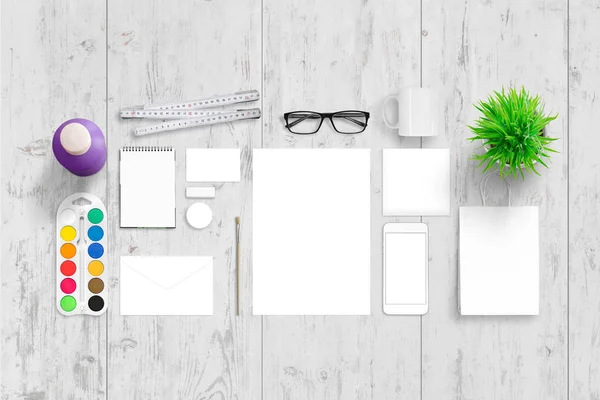Office stationery for corporate branding. Top view of white wooden desk with paper, pad, bag, envelope, business card, badge, mug, plant, glasses, water colors, brush and smart phone. — Stock Photo, Image