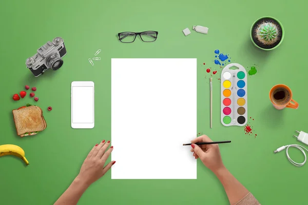 Flicka som målar på ett tomt vitt papper med borste och vatten färger. Ovanifrån av gröna skrivbord. — Stockfoto