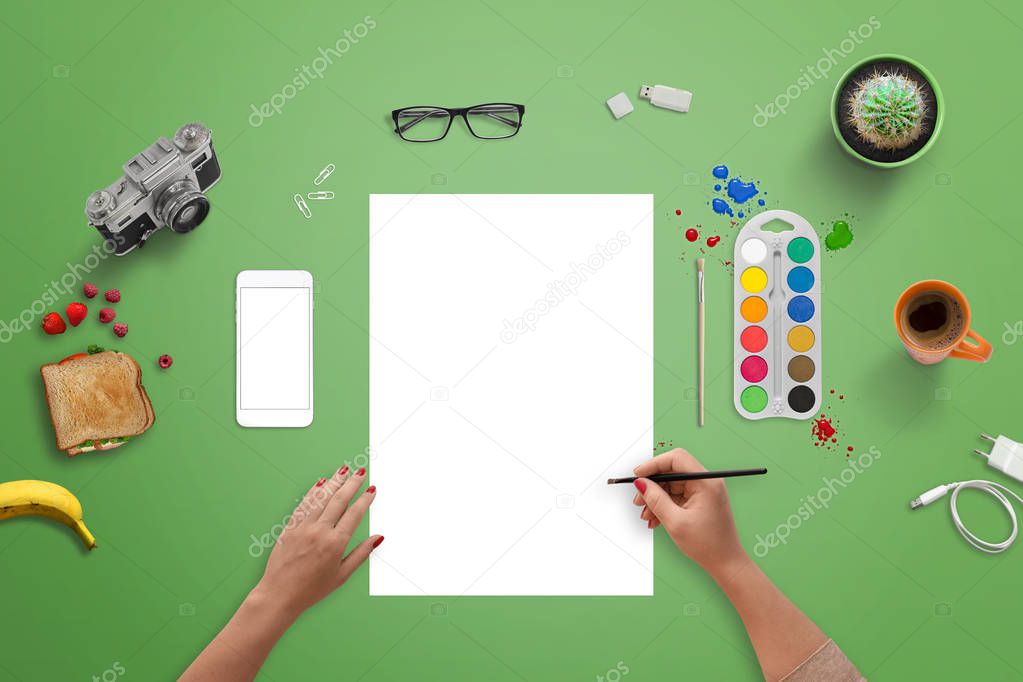 Girl painting on a blank white paper with brush and water colors. Top view of green desk.
