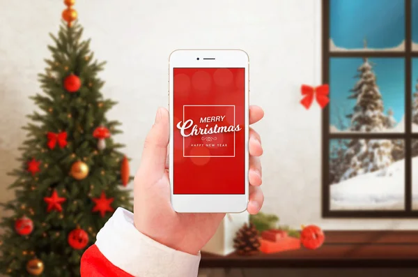 Teléfono móvil en la mano de Santa Claus mostrando Feliz Navidad y Feliz Año Nuevo saludo. Árbol de Navidad y decoraciones en el fondo — Foto de Stock