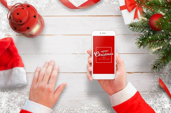 Santa con teléfono móvil en el escritorio. Árbol de Navidad con decoraciones, regalos y linterna en escritorio de madera blanca — Foto de Stock