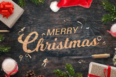 Neşeli Noel tebrik bir ahşap yüzey içine oyulmuş. Noel hediyeleri, ağaç dalları, keski, mumlar, pinecones ve Santa Claus şapka yanında.