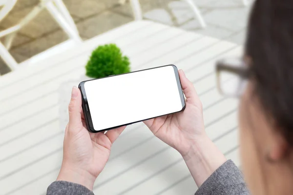 Woman holding smart phone in horizontal position. Isolated screen for mockup. White wooden desk and plant in background.