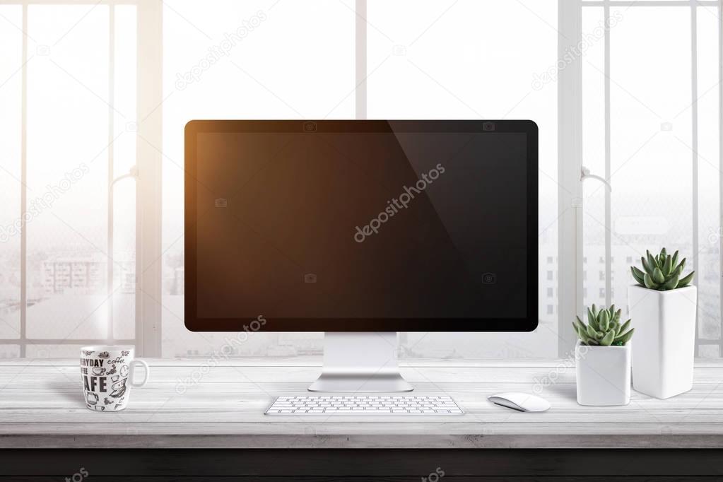 Computer display with blank screen for mockup in office or work room. Window and sun light in background. Cup of coffee and two plants beside.