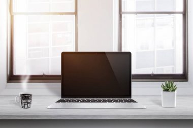Dizüstü bilgisayar ile fincan kahve ve yanında bitki ofis masasında. Güneş arka planda pencereden girer.