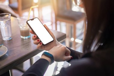 Akıllı telefon ve akıllı bilezik izleyen bir kadın. Model için boş ekranlı bilye. Arka planda kahve var.