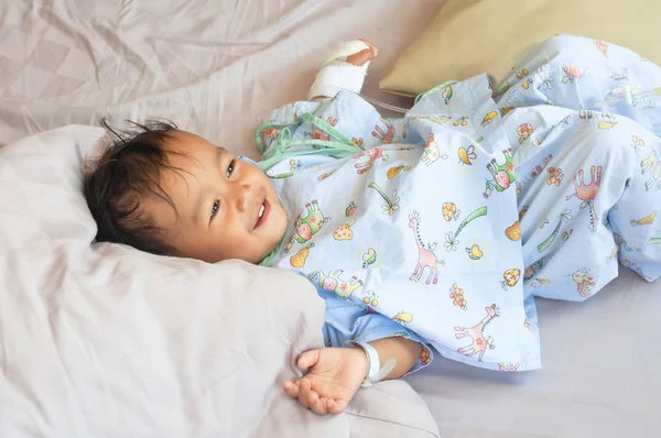 O rapazinho a olhar para os pais na cama, no quarto do hospital. Criança com tubo intravenoso. recuperação da doença . Imagem De Stock