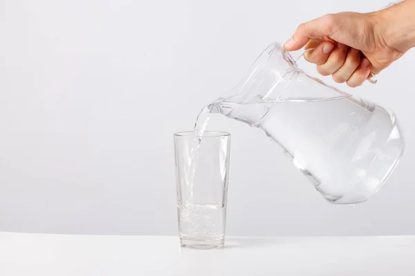 Рука наливает воду из стеклянного кувшина в стекло на белом фоне — стоковое фото