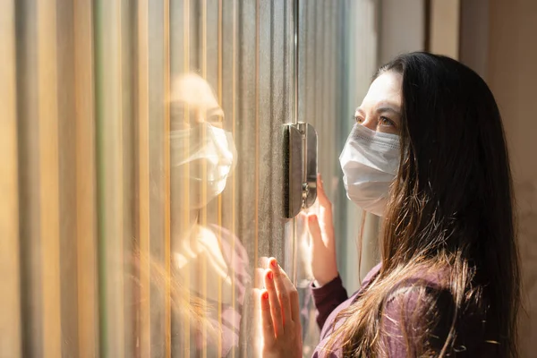 一名妇女在家中被隔离 戴着医疗面罩 身穿休闲装 呆呆地站在一扇关着的窗户前 手放在玻璃窗上 凝视着天空 待在家里的概念 — 图库照片