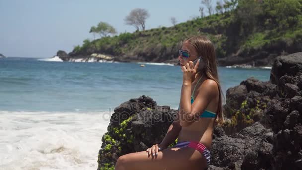 Chica con el teléfono móvil se sienta en la roca y mira al mar. Bali, Indonesia — Vídeos de Stock