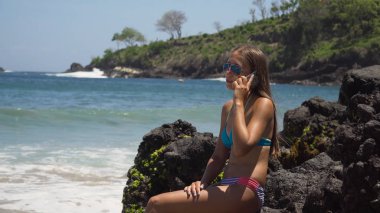 Cep telefonu olan kız kayanın üzerinde oturup denize bakıyor. Bali, Endonezya
