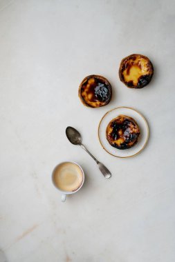 Pastel de nata. Geleneksel Portekiz tatlısı, seramik tabakta yumurtalı tart ve mermer arka planda bir fincan kahve ve kaşık. Üst görünüm