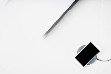 Kablosuz şarjda beyaz akıllı telefon ve beyaz arkaplanda hafif açık laptop. Üst Manzara. Boşluğu kopyala.