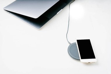 Kablosuz şarjda beyaz akıllı telefon ve beyaz arkaplanda hafif açık laptop. Yüksek açı. Boşluğu kopyala.