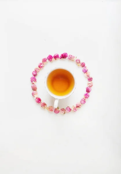 Cup of herbal tea over white marble background decorated with dry globe amaranth flowers. Circle pattern. Top view. Copy space.