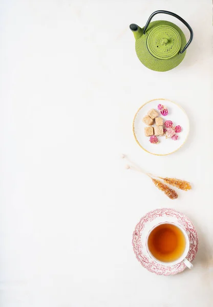 Cup of herbal tea and traditional oriental teapot over white marble background served with sugar cubes and dry globe amaranth flowers on a saucer and candy brown sugar on a stick. Top view. Copy space.