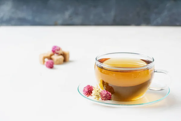 Cup of herbal tea over white marble background decorated with dried globe amaranth flowers, greec mountain tea and brown sugar cubes. Side view. Selective focus.