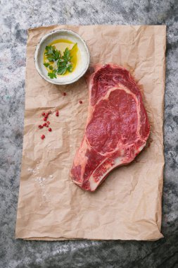 Kemiğin üzerinde çiğ biftek yeşillikler, tuz, baharatlar ve kırsal metal arka planda seramik bir kase zeytinyağı olan bir fırın kağıdı var. Üst görünüm