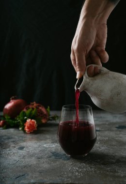 Erkek eli bardağa nar suyu döküyor. Nar meyveleri, tam ve yarım, arka planda nar çiçeği dallarıyla süslenmiş. Seçici odak