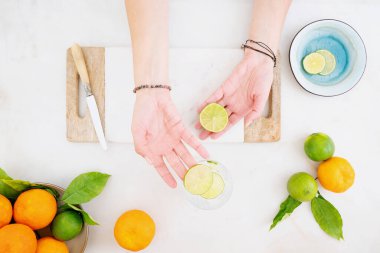 Citrus, su hazırladı. Dişi eller limon dilimlerini narenciye katılmış buzlu suya koyuyor. Masada taze portakal ve limon var. Üst görünüm.