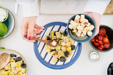 Geleneksel İtalyan makarna salatası. Orecchiette Arlecchino, kızarmış domates, soğan, siyah zeytin, limon, peynir, zeytinyağı, baharat ve otlar. Yemek pişirme süreci. Şef Feta peyniri ekliyor. Üst görünüm. 