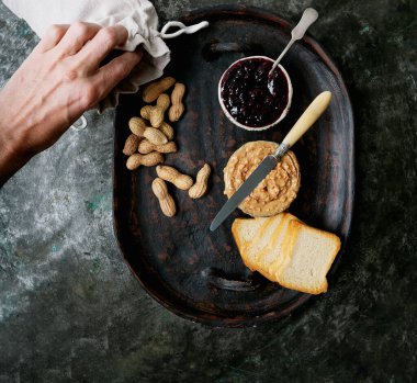 Fıstık ezmesi, ahududu reçeli ve kırsal metal arka planda tepside kızarmış ekmek dilimleri. Üst Manzara. Kahvaltı sandviçi hazırlığı. Erkek eli tepsiye fıstık döküyor..