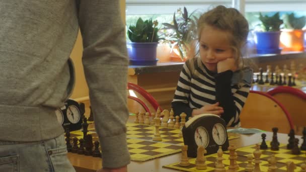 Gelangweilte blonde Mädchen spielen Schach beim Schachturnier für Vorschulkinder Organisator "Schwarzer Ritter" Kinder sitzen beim Schachbrettspiel im Klassenzimmer — Stockvideo