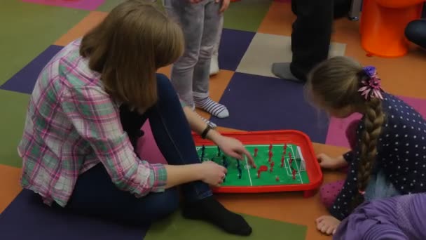 Niño y maestro están moviendo figuras jugando al futbol de mesa sentados en un piso colorido Los niños están alrededor de un niño rubio con trenza larga y educador — Vídeo de stock