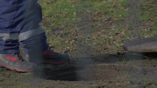 Kapanış aşağı bir yeraltı Hatch adamın ayak Boots erkekte kapanıyor bir Metal kap kullanarak bir demir sopa kanca ile onun son çamur çim güneşli günde çalışıyor — Stok video