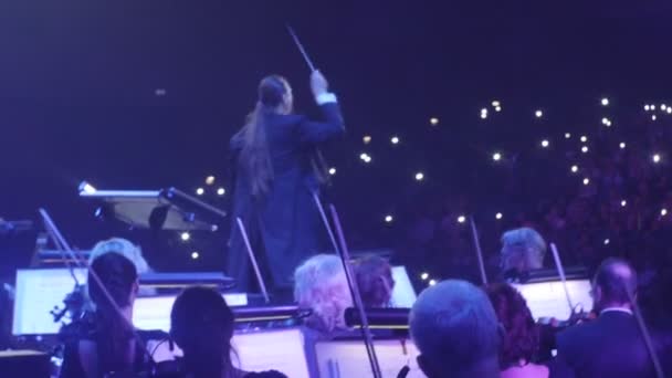 Rock symfoni konsert Kiev lång Haired dirigent Nikolai Lysenko är dirigering i främre av nyhetsradio musiker är spelar med ett bågar musikböcker på stativ — Stockvideo