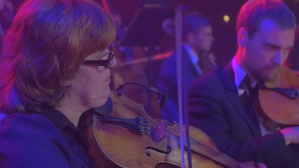 Rock Symphony Concert Kijów kobiet skrzypek gra skrzypce i uśmiechnięte ma przyjemność grupa muzyków grają niebieskie lampy oświetlenia ciemności Hall — Wideo stockowe