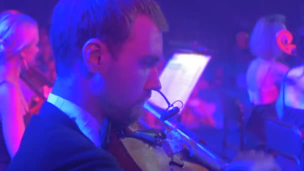 Ροκ συναυλία Συμφωνικής Κίεβο καλοντυμένος βιολιστές παίζουν σοβαρό αρσενικό μουσικός γυναίκες μουσικά βιβλία στην περίπτερα μπλε λαμπτήρες φωτισμού σκοτεινή αίθουσα τηλεθεατές — Αρχείο Βίντεο