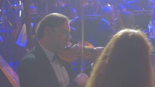 Szimfonikus koncert Kijevben rockegyüttes játszik hegedűre és hárfa zenét könyvek áll kék lámpa megvilágítás sötét terem férfi hegedűművész, előtérben a zenészek — Stock videók
