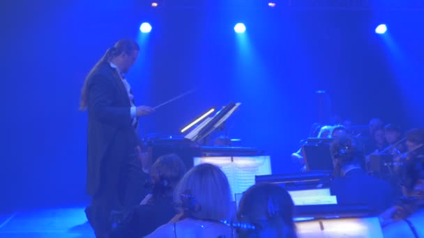 Rock Symphony koncert Kijów długowłosy mężczyzna zaczyna się do prowadzenia orkiestry macha z Baton muzycy grają niebieskie światła wielką orkiestrę powtarzania — Wideo stockowe