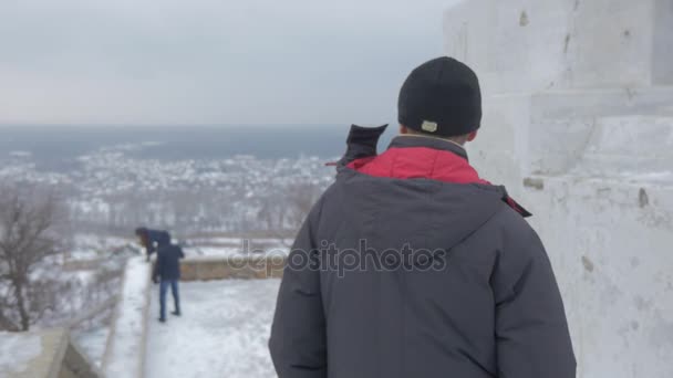 Um homem barbudo e dois jovens no convés de observação do monumento de Artyom com uma vista impressionante em Sviatogorskaya Lavra no tempo ventoso no inverno — Vídeo de Stock