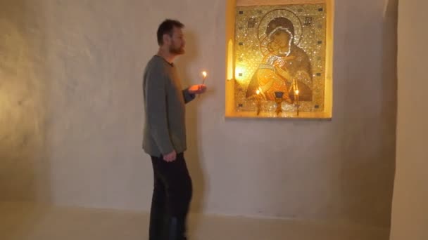 Un homme barbu debout de profil devant l'icône sainte et se dirigeant le long d'un étroit corridor blanc vers une croix orthodoxe jaune dans le temple des grottes de craie — Video