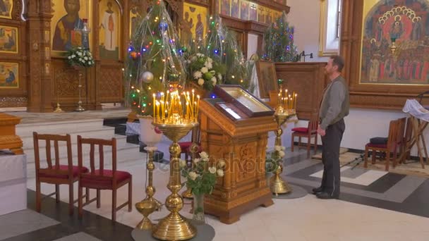 Um homem de meia-idade está diante de uma iconostase, que parece majestosa, com muitas velas de lit antes, árvores de abeto e ícones de aparência dourada — Vídeo de Stock