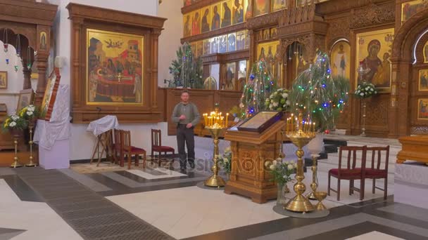 Prachtvolle christliche Ikonostase mit einem bärtigen Mann davor, der Tannenbäume, Blumen, golden aussehende Ikonen betrachtet und anfängt zu beten — Stockvideo